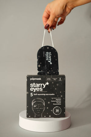 Hand holding a black Starry Eyes Warming Eye Mask - Single | Popmask with a star pattern above a matching box containing five self-warming eye masks for wellness and skincare.