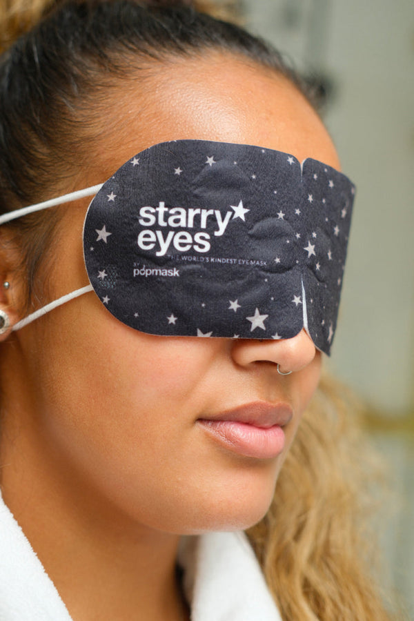 A woman wearing the Starry Eyes Warming Eye Mask - Single | Popmask, a black eye mask with white star patterns designed for skincare and wellness relaxation.