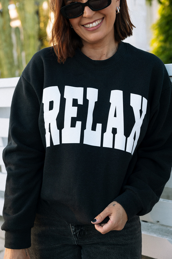 Woman wearing black RELAX Collegiate Crew Neck Sweatshirt | Lucky Owl with bold white letters, paired with dark jeans and sunglasses, smiling outdoors in natural light.