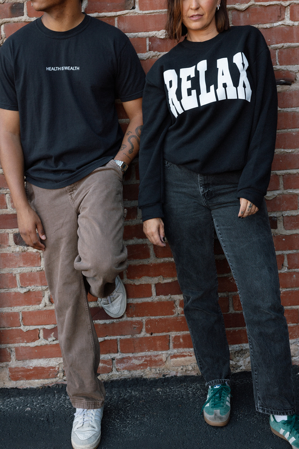 A person wearing the RELAX Collegiate Crew Neck Sweatshirt | Lucky Owl in black with white lettering, standing next to another person in casual clothing against a brick wall.