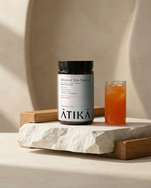 Jar of Advanced Skin Nutrition | ATIKA dietary supplement next to a glass of iced beverage on a textured stone and wooden display, promoting skincare and wellness supplements.