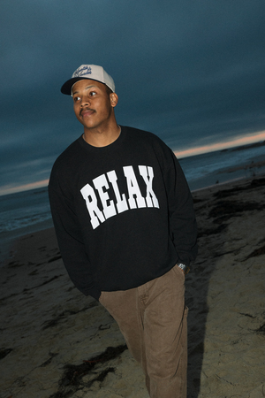 Man wearing a black RELAX Collegiate Crew Neck Sweatshirt | Lucky Owl with bold white lettering, standing on a beach at dusk with a calm ocean background.