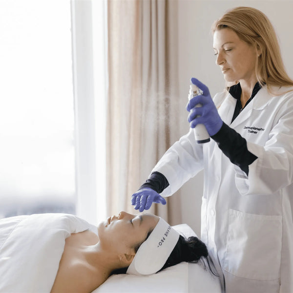A skincare professional wearing gloves sprays Satisfaction Mist | DermaplanePro on a relaxed woman's face in a bright, calming treatment room.