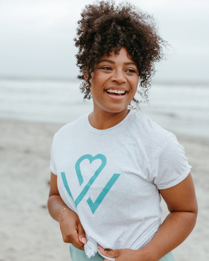 Smiling woman with curly hair standing on a beach wearing a white Wellness Month Logo – Unisex Shirt | Wellness Month featuring a teal heart and checkmark design.