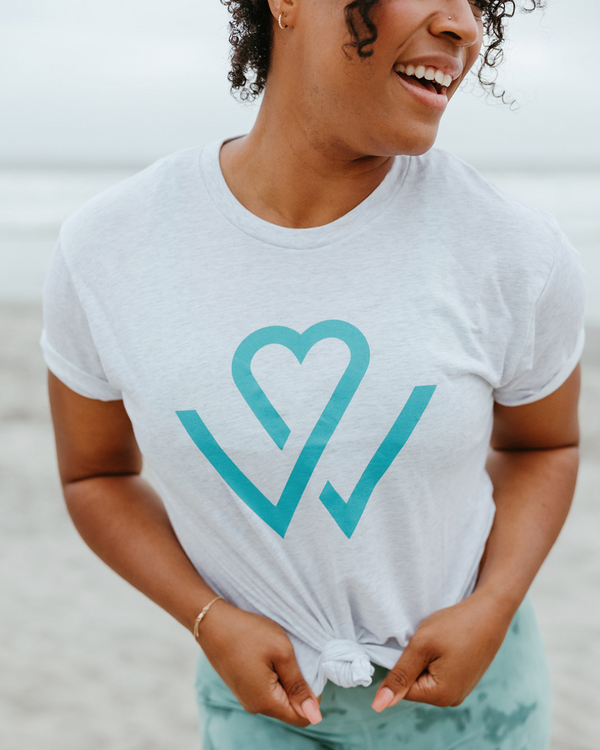 Person smiling and wearing a white Wellness Month Logo – Unisex Shirt | Wellness Month with a turquoise heart-shaped design, promoting wellness and positive energy.