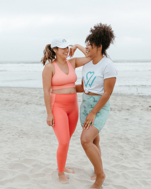 Two women smiling and chatting on a beach, one wearing a Wellness Month Logo – Unisex Shirt | Wellness Month and the other in coral activewear, showcasing a joyful outdoor wellness moment.