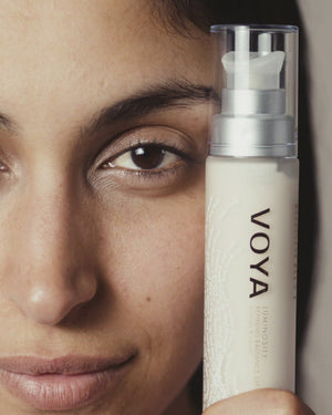 Close-up of a woman holding a bottle of Luminosity Refining Radiance Exfoliator | VOYA near her face, showcasing natural skincare and wellness.