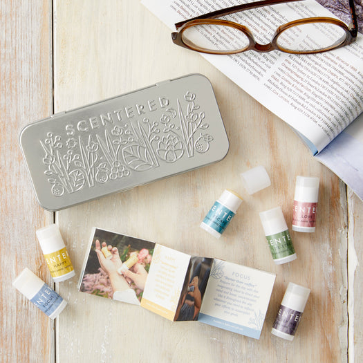 Wellbeing Ritual Aromatherapy Mini Tin | Scentered with six small aromatherapy balm sticks in various colors, accompanied by a booklet and reading glasses on a light wooden surface.