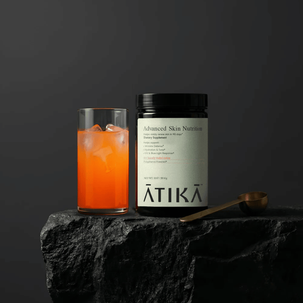 Jar of Advanced Skin Nutrition | ATIKA dietary supplement next to a glass of orange liquid with ice, placed on a textured black stone surface with a gold spoon nearby