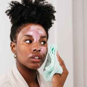 Woman with natural hair applying a pink and white skincare mask on her face while holding a soft light blue towel, showcasing the Moon Silk Moisture Recharging Mask | COOLA in a self-care routine.