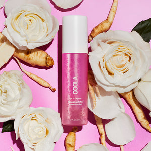 Bottle of Rewakening™ Rosewater Mist Face Spray | COOLA surrounded by white rose flowers and root ingredients on a pink background, highlighting skincare and natural wellness.