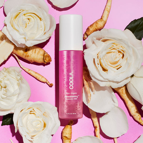 Bottle of Rewakening™ Rosewater Mist Face Spray | COOLA surrounded by white rose flowers and root ingredients on a pink background, highlighting skincare and natural wellness.