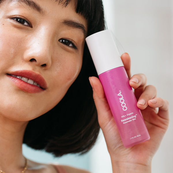 A woman holding a pink bottle of Rewakening™ Rosewater Mist Face Spray | COOLA close to her face, showcasing clear, glowing skin in a bright setting.