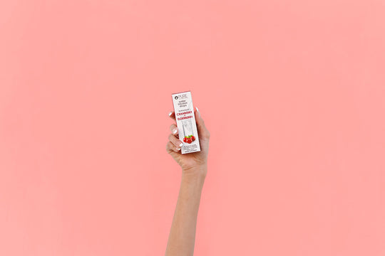 Hand holding a box of Super Fruit Extracts Water Infusion Drops - Cranberry + Elderberry | Pure Inventions against a soft pink background, highlighting wellness supplement packaging.
