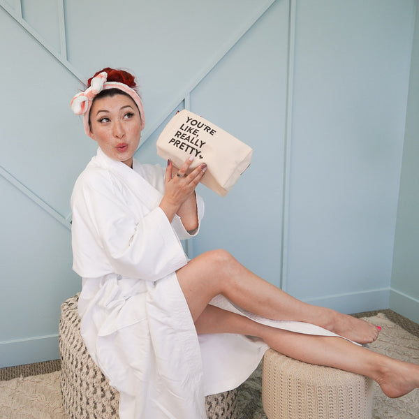 A woman in a white robe and pink headband holds the "You're Like, Really Pretty Bag | Live Love Spa" while sitting on a woven ottoman in a stylish, light-colored room.