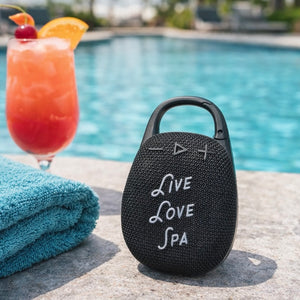 Wireless Clip Speaker | Live Love Spa in black with textured surface, placed by a pool next to a blue towel and a tropical drink, showcasing a portable wellness device for outdoor use.