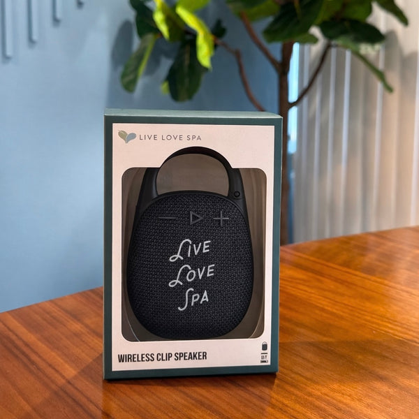 Wireless Clip Speaker | Live Love Spa in black with textured surface, featuring control buttons and branded text, displayed in branded packaging on a wooden table with green plant background.