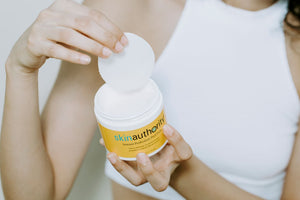 Hands holding a jar of Instant Perfection Peel Pads | Skin Authority with a white peel pad being lifted from the container for skincare use.