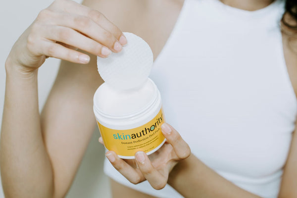 Hands holding a jar of Instant Perfection Peel Pads | Skin Authority with a white peel pad being lifted from the container for skincare use.