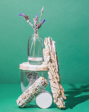 Clear glass jar with dried lavender flowers next to bundles of Palo Santo and sage, evoking natural wellness and relaxation, featuring Palo Santo Sage Rollerball Perfume Oil - Spirit | Hydra Bloom.