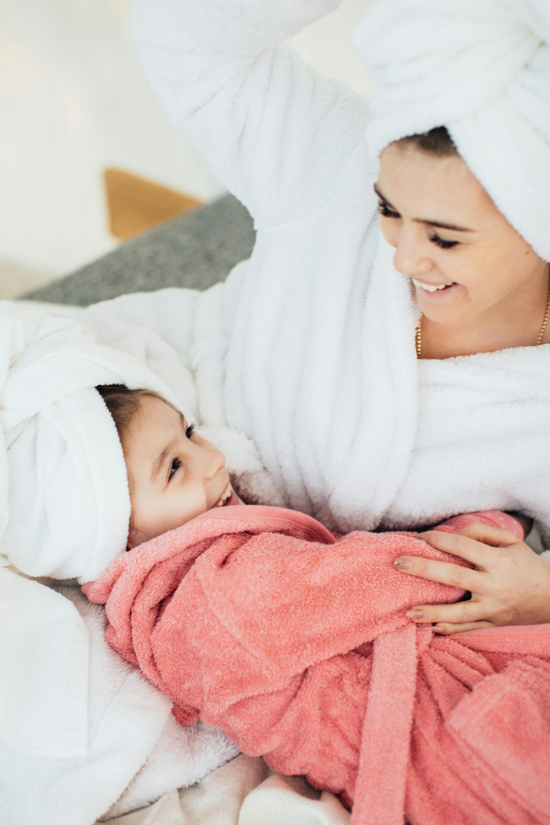 A smiling woman and young girl wearing white and pink robes with matching white towels wrapped around their heads, sharing a joyful moment in a cozy setting.
