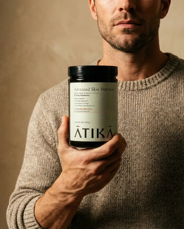 Man in a beige sweater holding a container of Advanced Skin Nutrition | ATIKA supplement for skincare and wellness, against a neutral background.