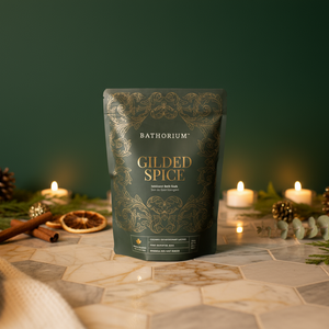 Gilded Spice Limited Edition Bath Soak | Bathorium in elegant packaging surrounded by warm candles, cinnamon sticks, dried orange, and pinecones for a cozy spa and wellness setting.