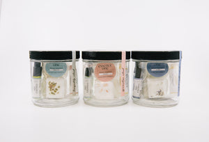 Three clear jars with black lids, containing small vials, dried herbs, and labeled items, showcasing the Ritual Kit | Essence One for skincare and wellness.