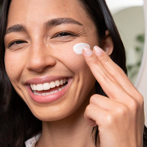 Young woman smiling while applying Dew Good Illuminating Serum Probiotic Sunscreen SPF 30 | COOLA to her cheek in a bright skincare routine moment.