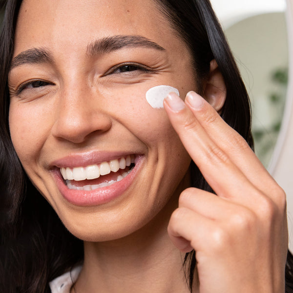 Young woman smiling while applying Dew Good Illuminating Serum Probiotic Sunscreen SPF 30 | COOLA to her cheek in a bright skincare routine moment.