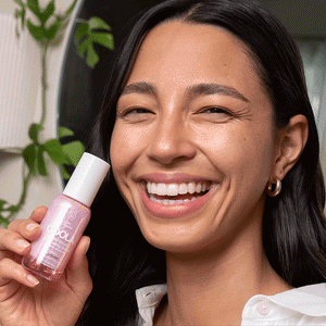 Smiling woman holding Dew Good Illuminating Serum Probiotic Sunscreen SPF 30 | COOLA bottle, highlighting skincare and natural glowing skin in a bright indoor setting.