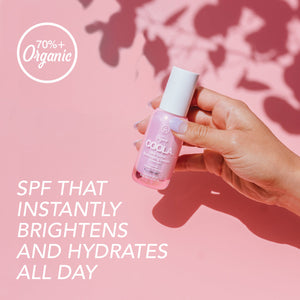 Hand holding a small pink bottle of Dew Good Illuminating Serum Probiotic Sunscreen SPF 30 | COOLA against a pink background with text highlighting its brightening and hydrating benefits and 70%+ organic ingredients.