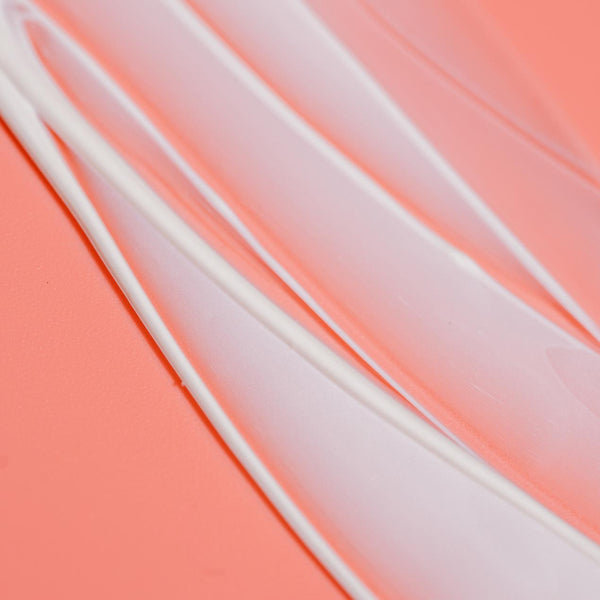 Close-up texture of smooth white serum spread on a soft coral background, representing Dew Good Illuminating Serum Probiotic Sunscreen SPF 30 | COOLA skincare product.