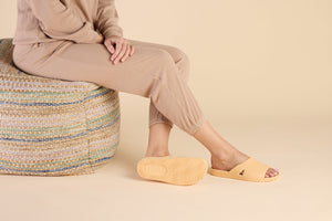 Person sitting on a woven pouf wearing stylish tan slip-on sandals with textured soles, showcasing Bonsai Slides Tan | Feelgoodz in a cozy, neutral setting.
