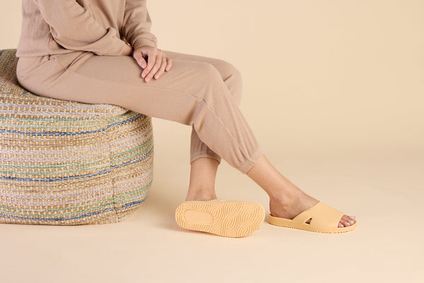 Person sitting on a woven pouf wearing stylish tan slip-on sandals with textured soles, showcasing Bonsai Slides Tan | Feelgoodz in a cozy, neutral setting.
