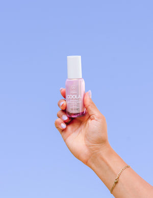 Hand holding a pink bottle of Dew Good Illuminating Serum Probiotic Sunscreen SPF 30 | COOLA against a clear blue sky, showcasing skincare in a wellness context.