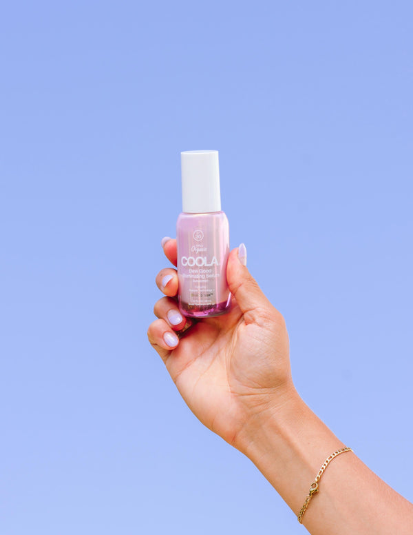 Hand holding a pink bottle of Dew Good Illuminating Serum Probiotic Sunscreen SPF 30 | COOLA against a clear blue sky, showcasing skincare in a wellness context.