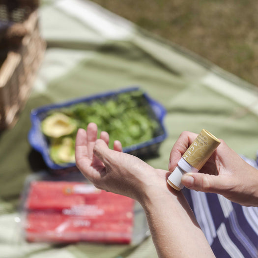 Person applying Be Happy Mini Aromatherapy Balm | Scentered on their wrist outdoors with a picnic basket and fresh vegetables in the background, suggesting a moment of wellness and self-care.