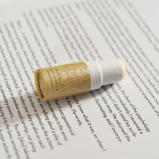 Be Happy Mini Aromatherapy Balm | Scentered in a small yellow container resting on an open book page, promoting wellness and skincare with grapefruit and spearmint scents.