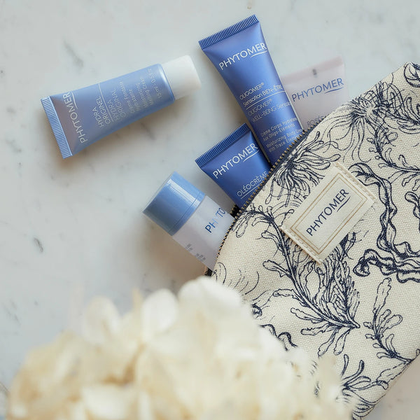 Compact cosmetic tubes and containers from the Seaside Moisturizing Starter Kit | Phytomer arranged next to a textured toiletry bag with botanical design on a marble surface.