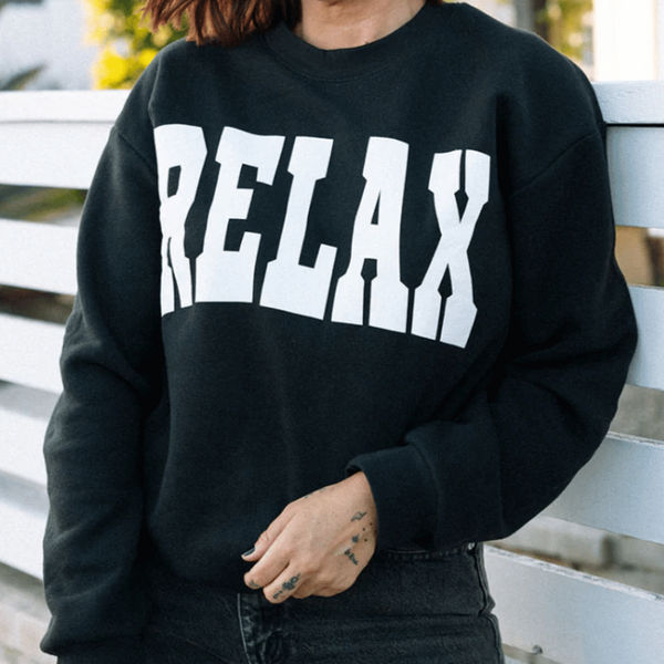 Person wearing a black RELAX Collegiate Crew Neck Sweatshirt | Lucky Owl with bold white lettering, standing outdoors near a white fence in casual wear.