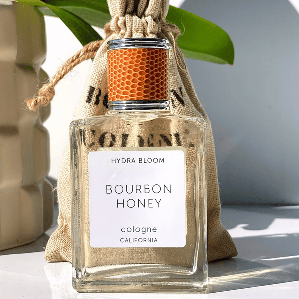 Clear glass bottle of Bourbon + Honey Cologne - Unisex Masculine Scent | Hydra Bloom with textured honeycomb-patterned cap, natural light, and burlap pouch in background.