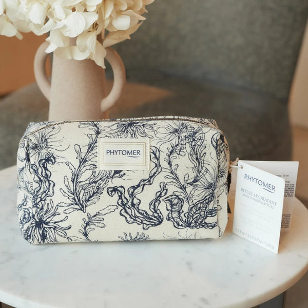 Seaside Moisturizing Starter Kit | Phytomer in a cream-colored pouch with dark blue seaweed pattern, displayed on a marble table with a beige vase of white flowers.