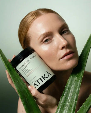 Woman with clear skin holding a jar of Advanced Skin Nutrition | ATIKA skincare product next to fresh aloe vera leaves, highlighting natural wellness and body care.