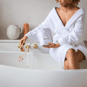 A person in a white bathrobe sprinkling Amber Glow Clay Mineral Soak | Bathorium into a bathtub filled with water, evoking a relaxing skincare and wellness spa experience.