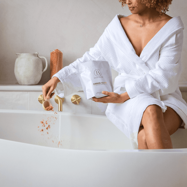 A person in a white bathrobe sprinkling Amber Glow Clay Mineral Soak | Bathorium into a bathtub filled with water, evoking a relaxing skincare and wellness spa experience.