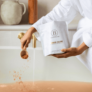 Person in a white robe holding a package of Amber Glow Clay Mineral Soak | Bathorium while pouring orange clay mineral soak into a bathtub filled with warm water for a relaxing skincare and body care experience.