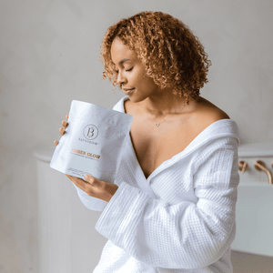 A woman in a white bathrobe holding and looking at a package of Amber Glow Clay Mineral Soak | Bathorium in a serene bathroom setting.