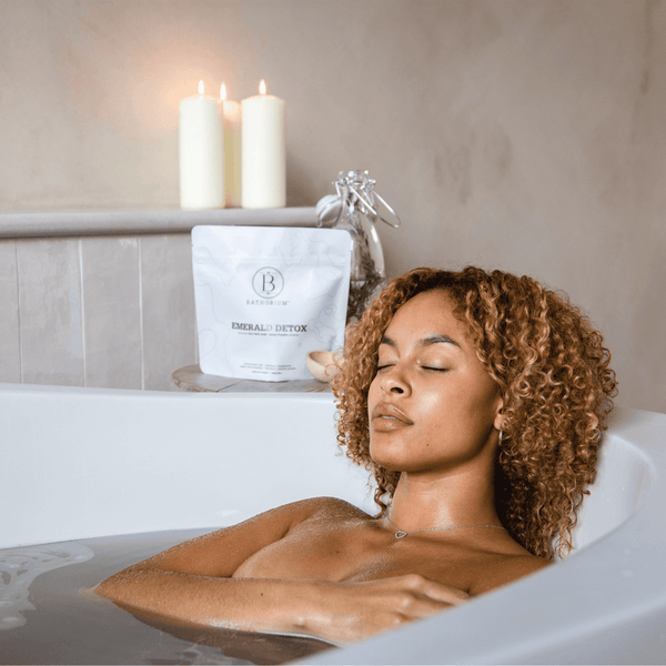 A woman relaxing in a bathtub with eyes closed, next to a package of Emerald Detox Clay Mineral Soak | Bathorium and lit candles creating a calm wellness atmosphere.