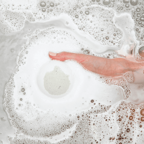 A hand gently touching bubbly water surrounding a dissolving bath bomb, showcasing the Boreal Fog Bath Bomb | Bathorium in a relaxing bath setting.
